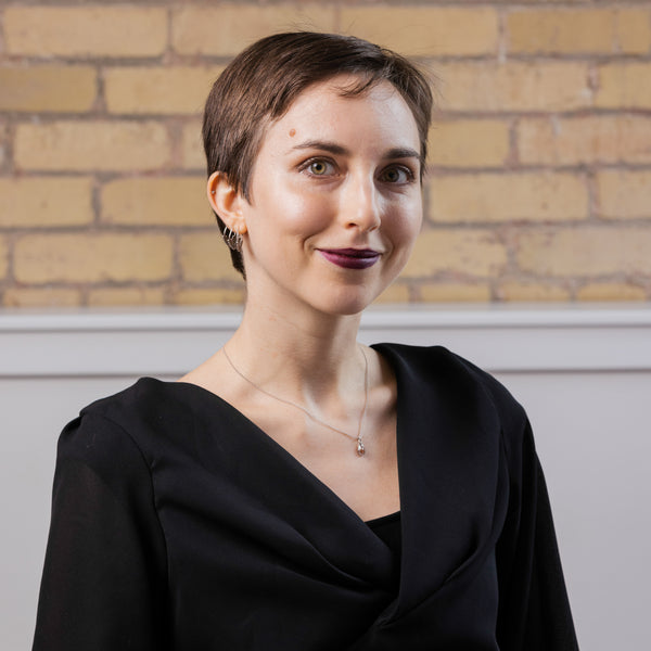 Person with short brown hair and purple lipstick wearing a black outfit against a brick wall background