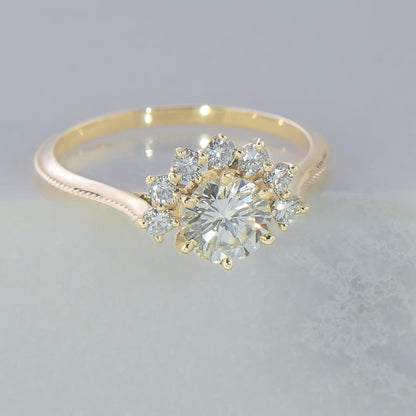 Handmade in 14 karat recycled yellow gold this cluster ring contains 7 recycled 1.75mm diamonds surrounding a 0.68 carat recycled round solitaire diamond. The shank has a graduated, cathedral profile with millgrain on the top profile.