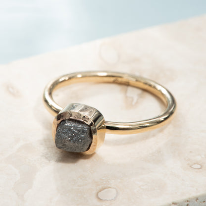 A rounded square bezel setting holding a grey rough diamond on a polished yellow gold band.