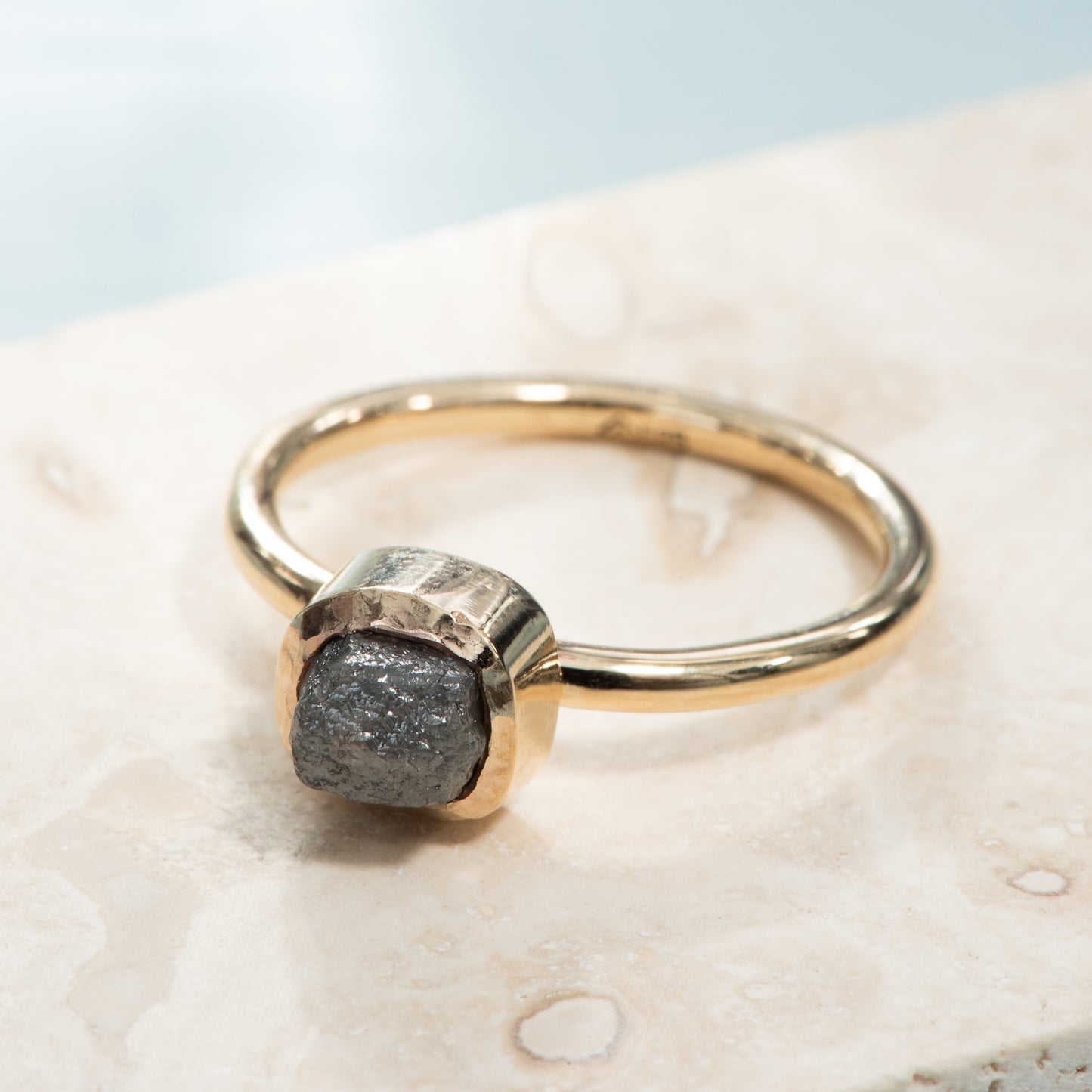 A rounded square bezel setting holding a grey rough diamond on a polished yellow gold band.