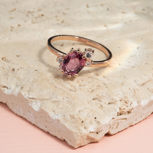 An oval cut pink sapphire is set on a slant on a polished rose gold band, with 5 small colourless diamonds asymmetrically set around it.