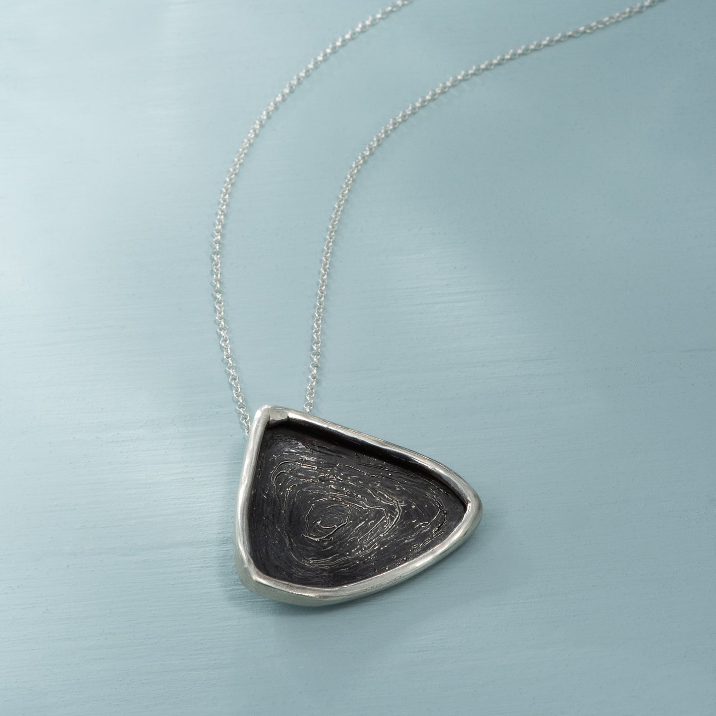 A large rounded triangular silver pendant, oxidized black with a thin ring of polished silver at the edges, on a long silver chain.