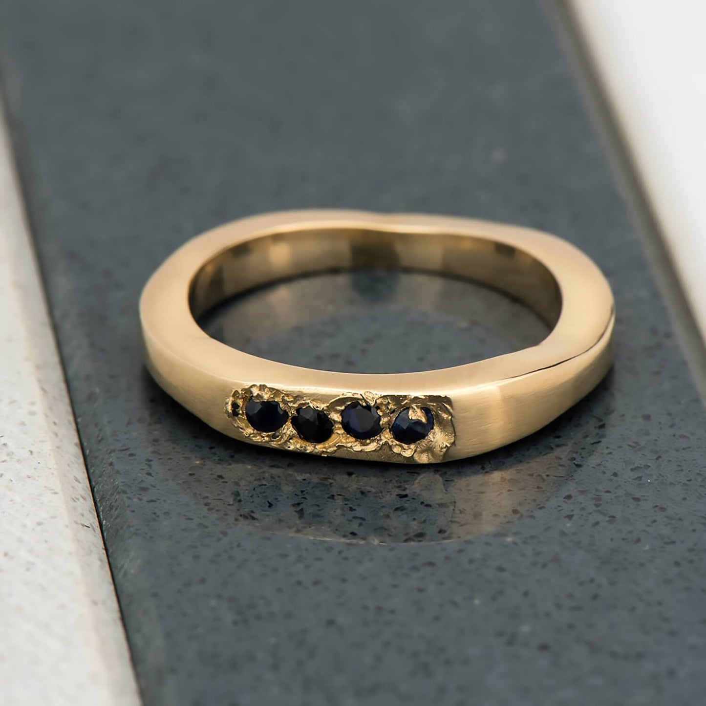 Recycled 14 karat yellow gold signet ring with four 2mm natural blue sapphires in which have been set in cast, casted. The surroundings of the stones have been finished with a rough baroque texture and the band is an irregular shape.